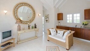 A spacious living area with a large round mirror, a console table, a sofa, and a kitchen area with wooden cabinets and a window. - Luxury 6 Bed Villa With Pool, Mullins Bay (Mullins)