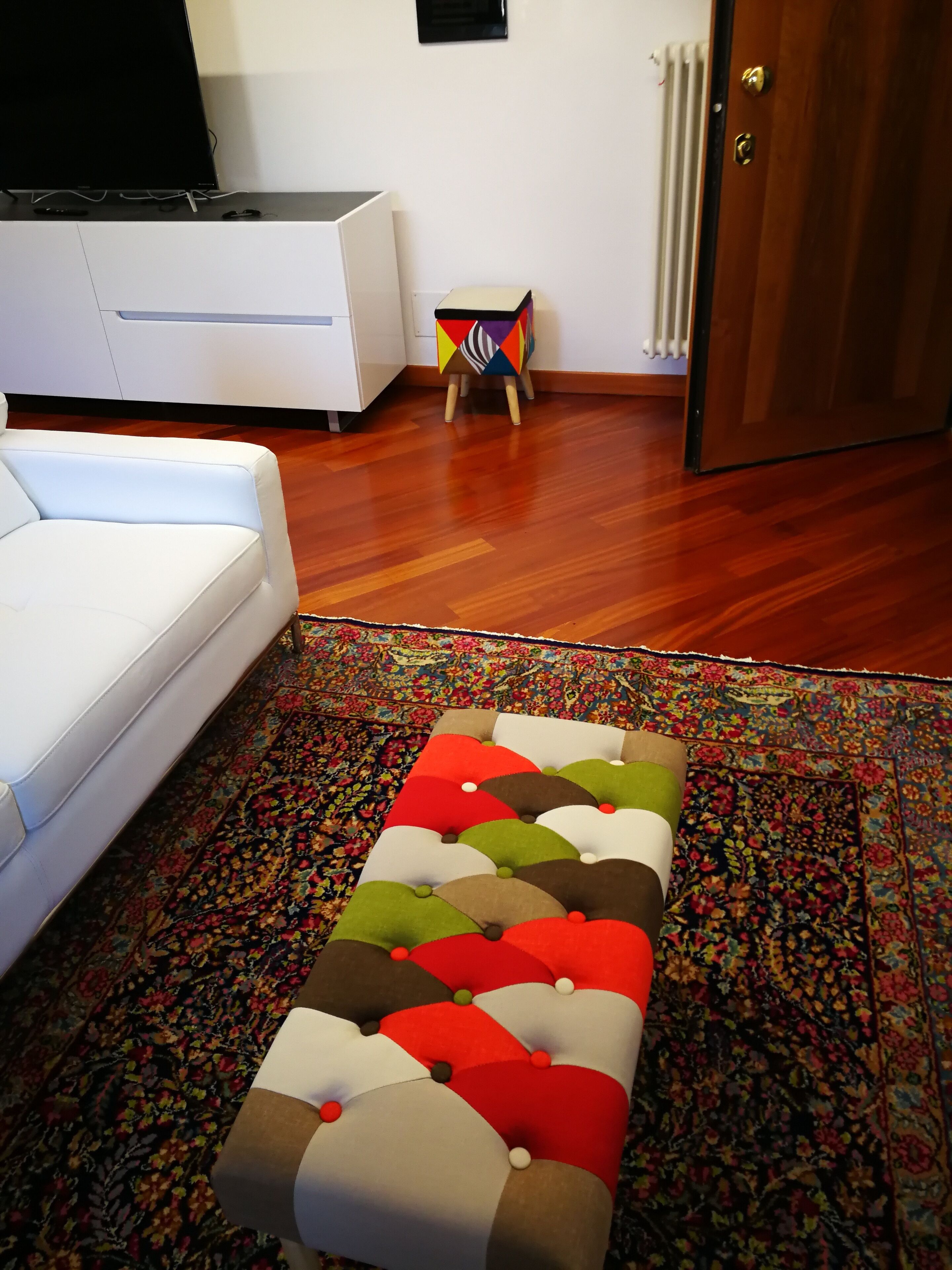 A living room with a white sofa, a colorful ottoman, a patterned rug, and a radiator.