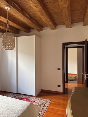 A room with a wooden ceiling, a white wardrobe, a hanging light fixture, a patterned rug, and a door leading to another room.