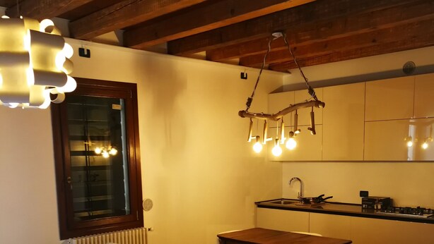 A modern living room with a white sofa, wooden ceiling beams, and a kitchen area in the background.