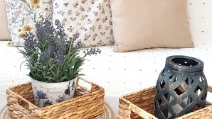 A white sofa with floral and solid cushions, two woven baskets with a plant and a black vase, and a woven placemat on a white coffee table.