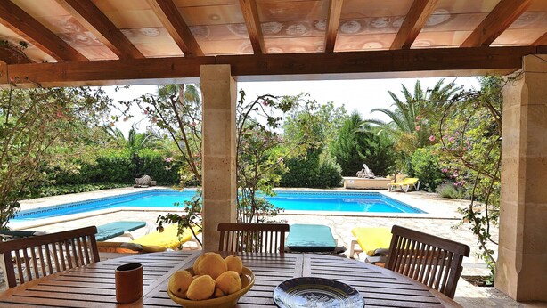 A wooden table with a bowl of lemons and a plate, set under a wooden pergola with a view of a swimming pool and lush greenery.