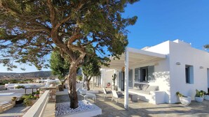 A modern white building with a stone patio, outdoor seating, and a large tree. - Villa Meraki (Agkeria)