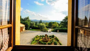 Vista a través de una ventana de un jardín con una fuente central, rodeado de arbustos y árboles cuidadosamente podados.