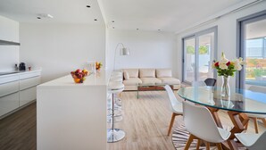 Een moderne keuken met witte kasten, een centraal eiland met barkrukken, een eetgedeelte met een glazen tafel en witte stoelen, en een beige bank op de achtergrond.