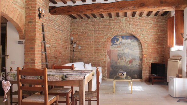 Une pièce rustique avec un mur en briques, des poutres en bois au plafond, un lit, une table avec des chaises, une petite table ornée d’un objet décoratif et un téléviseur.