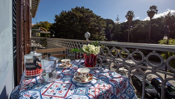 Un balcón con una mesa dispuesta para dos personas, con mantel estampado, una cafetera, tazas y un florero con flores.