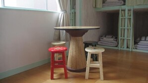 A room with a round wooden table surrounded by stools, a window with curtains, and a shelf with folded blankets. - Kichinan - Hostel (Osaka)