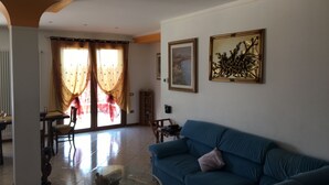 A living room with a blue sofa, a glass coffee table, and a dining area with a table and chairs. There are two windows with curtains, and a painting on the wall.