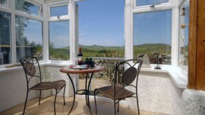 A room with two wicker chairs and a small round table, a bottle of wine, and a view of a green landscape through large windows. - Escape To This Hidden Gem & Relax In Tranquility - Beaches & Coastal Path Nearby (st nicholas)
