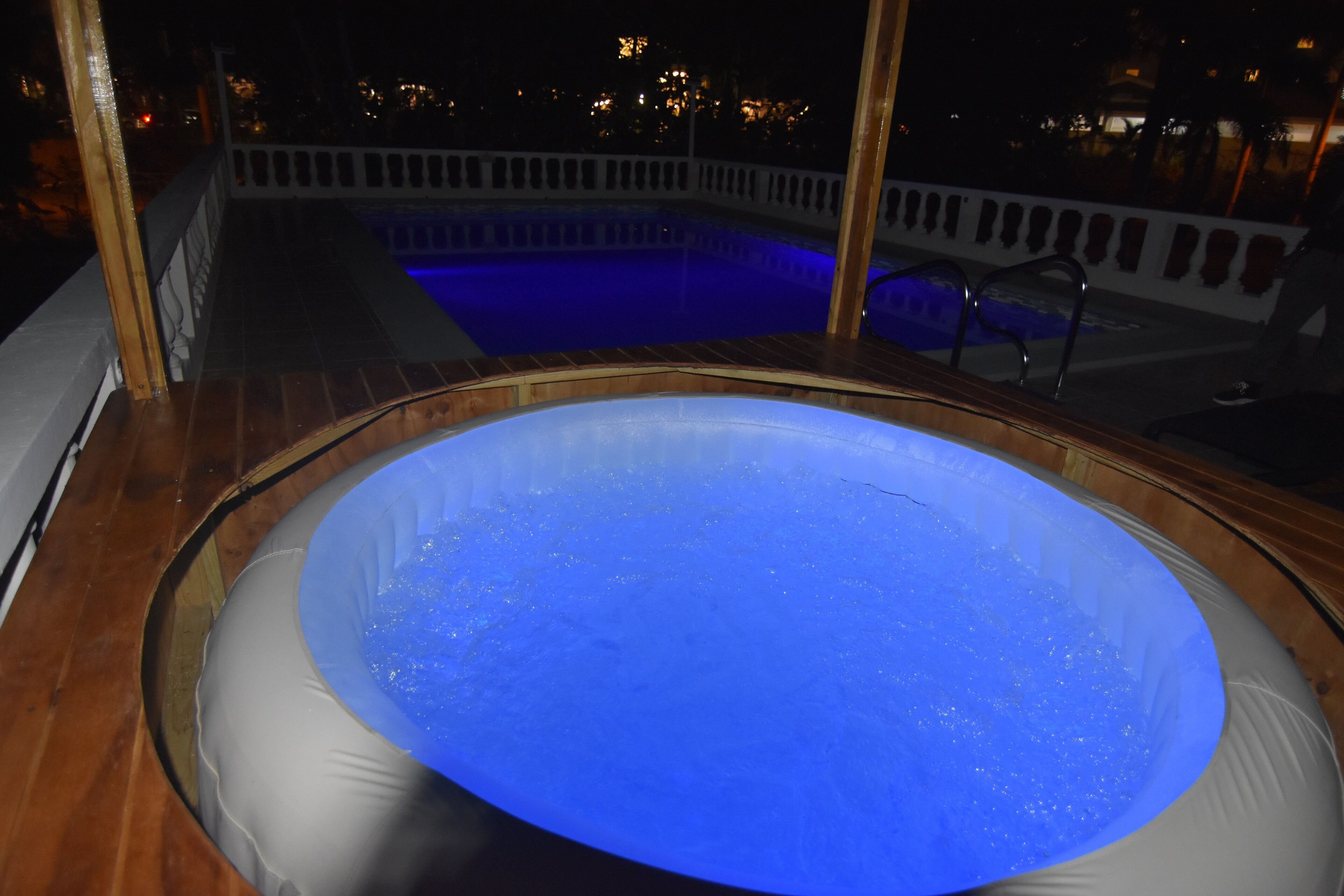 A hot tub with a blue light, surrounded by a wooden deck and a pool in the background.