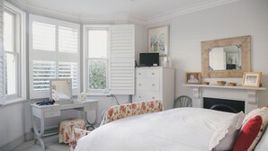 A bedroom with a bed, a desk, a chair, a fireplace, and a window with shutters.