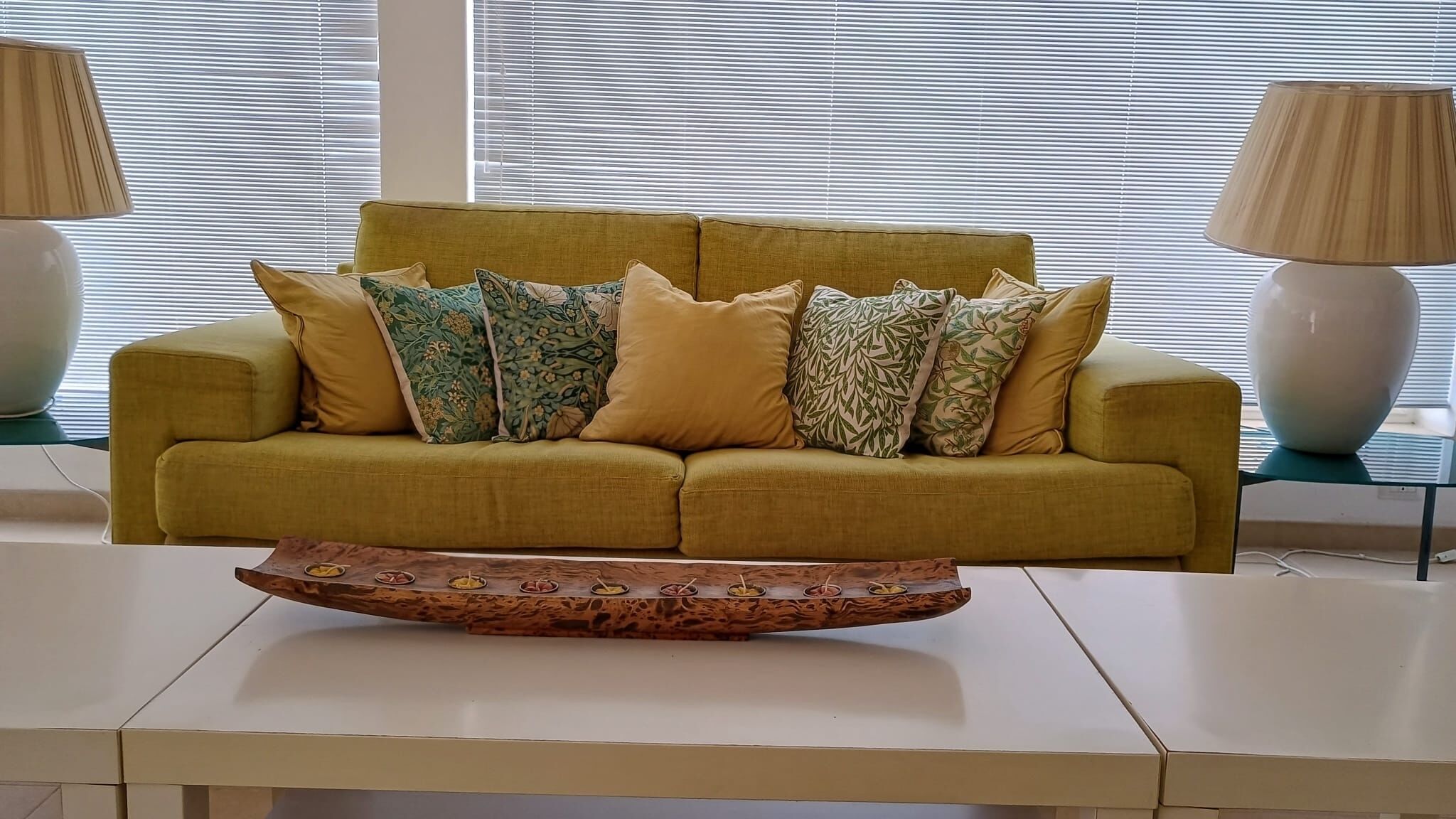 A mustard-colored sofa with patterned cushions, a wooden tray with fruit, and two lamps on either side.