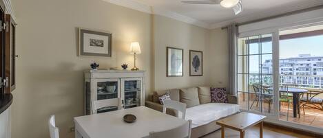 A living room with a white dining table, white chairs, a sofa, and a ceiling fan. There are framed pictures on the wall and a view of the outside through large windows.