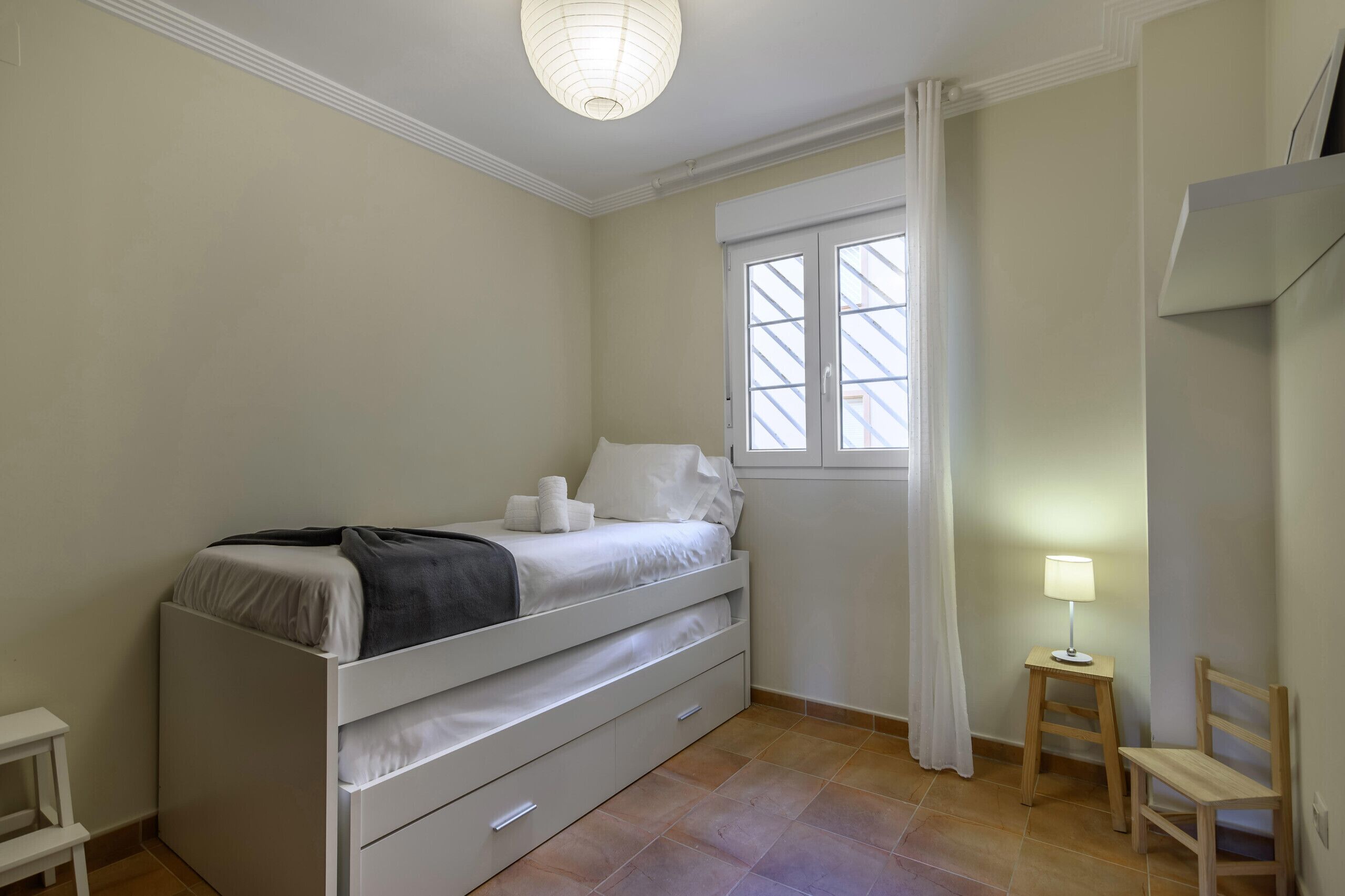 Ein Schlafzimmer mit einem Bett, einem kleinen Tisch mit einer Lampe, einem Sessel und einem Fenster mit Vorhängen.