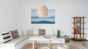 A modern living room with a white sofa, a round coffee table, a woven pendant light, a bookshelf, and a framed picture of the ocean.