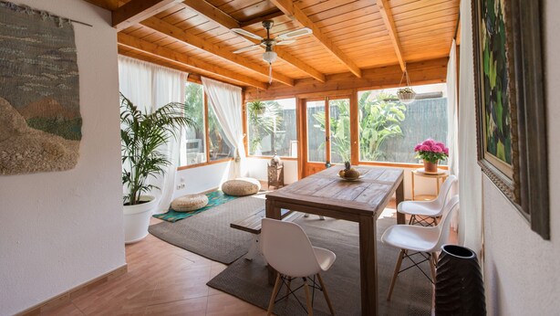 A spacious room with a wooden ceiling, a dining table with chairs, a large window with curtains, and a potted plant.