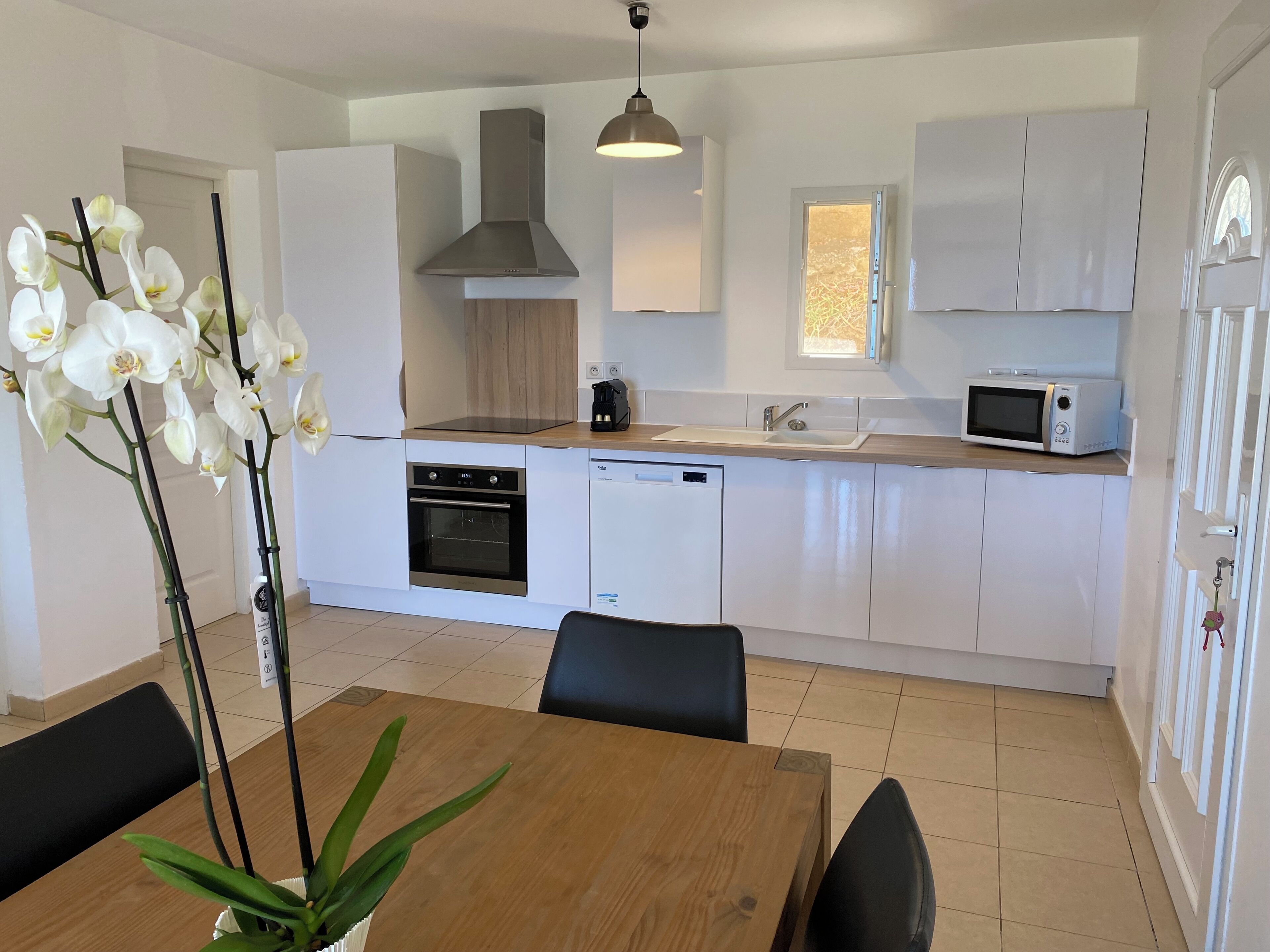 A modern kitchen with white cabinets, a built-in oven, a microwave, and a sink. There is a dining table with chairs and a potted orchid.