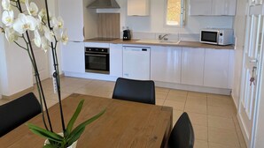 Une cuisine moderne avec des armoires blanches, un four encastré, un four à micro-ondes et un évier. On y trouve une table à manger avec des chaises et une orchidée en pot.
