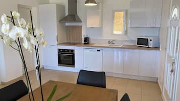 A modern kitchen with white cabinets, a built-in oven, a microwave, and a sink. There is a dining table with chairs and a potted orchid.