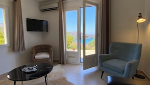 Une pièce avec un fauteuil en osier, un fauteuil bleu, une table noire avec une télécommande, et une vue sur la mer par une porte coulissante.