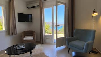 A room with a wicker chair, a blue armchair, a black table with a remote, and a view of the sea through a sliding door.