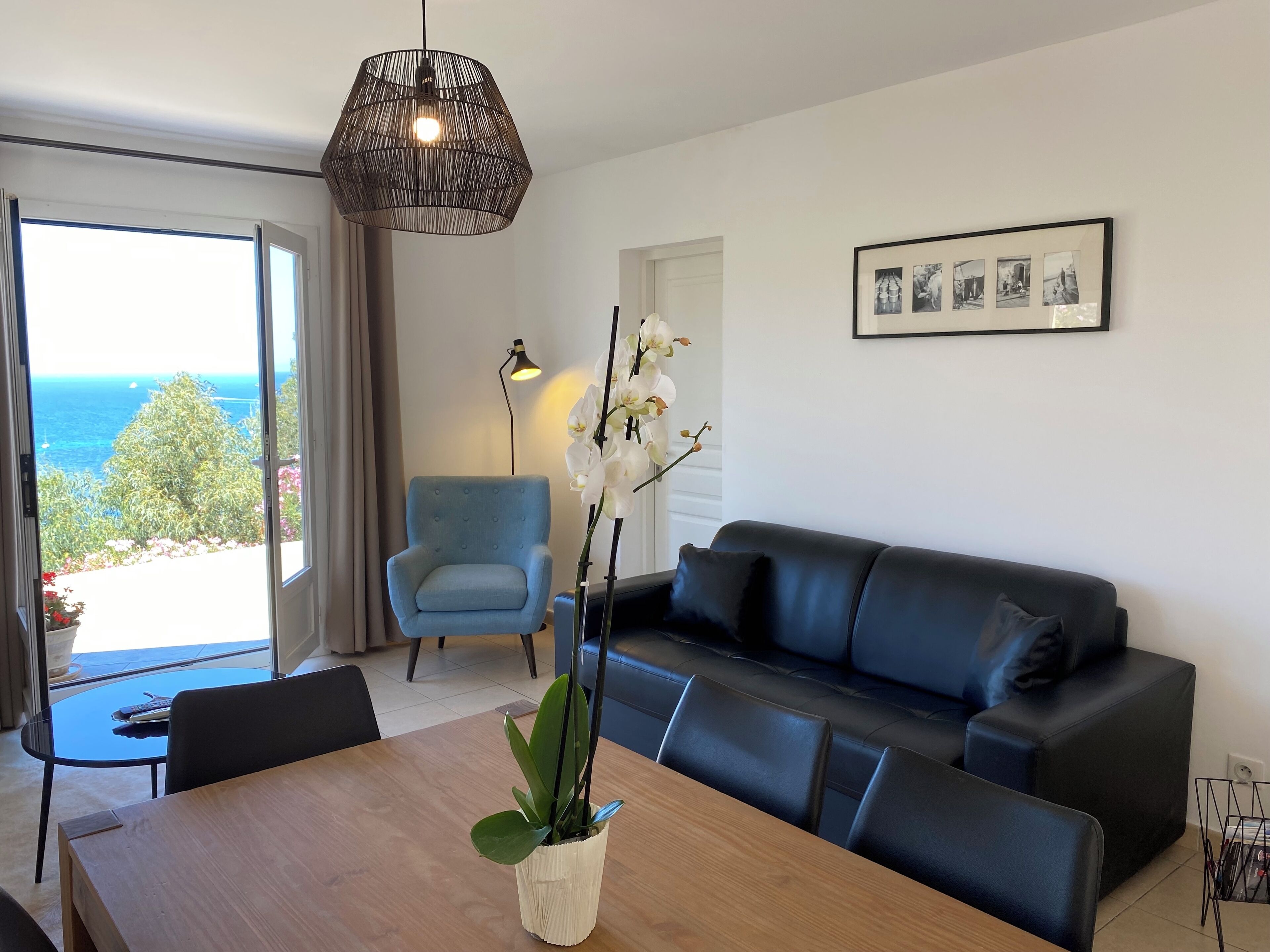 Ein modernes Wohnzimmer mit einer Couch, einem Esstisch mit Stühlen, einem blauen Sessel und einem großen Fenster mit Meerblick.