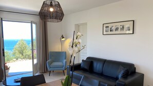 Un salon moderne avec un canapé, une table à manger et des chaises, un fauteuil bleu et une grande fenêtre donnant sur la mer.