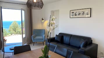 Ein modernes Wohnzimmer mit einer Couch, einem Esstisch mit Stühlen, einem blauen Sessel und einem großen Fenster mit Meerblick.