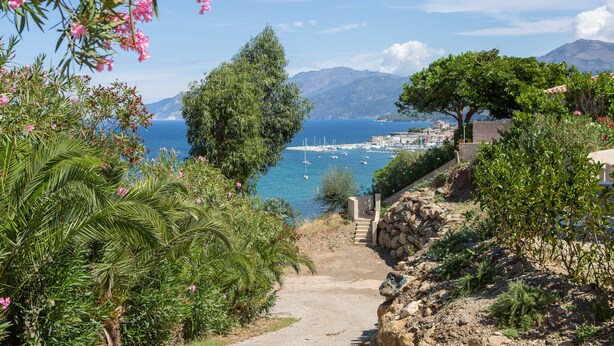 Un sentier côtier mène à une marina où sont amarrés des bateaux, le tout étant entouré d’une végétation luxuriante et avec des montagnes en toile de fond.