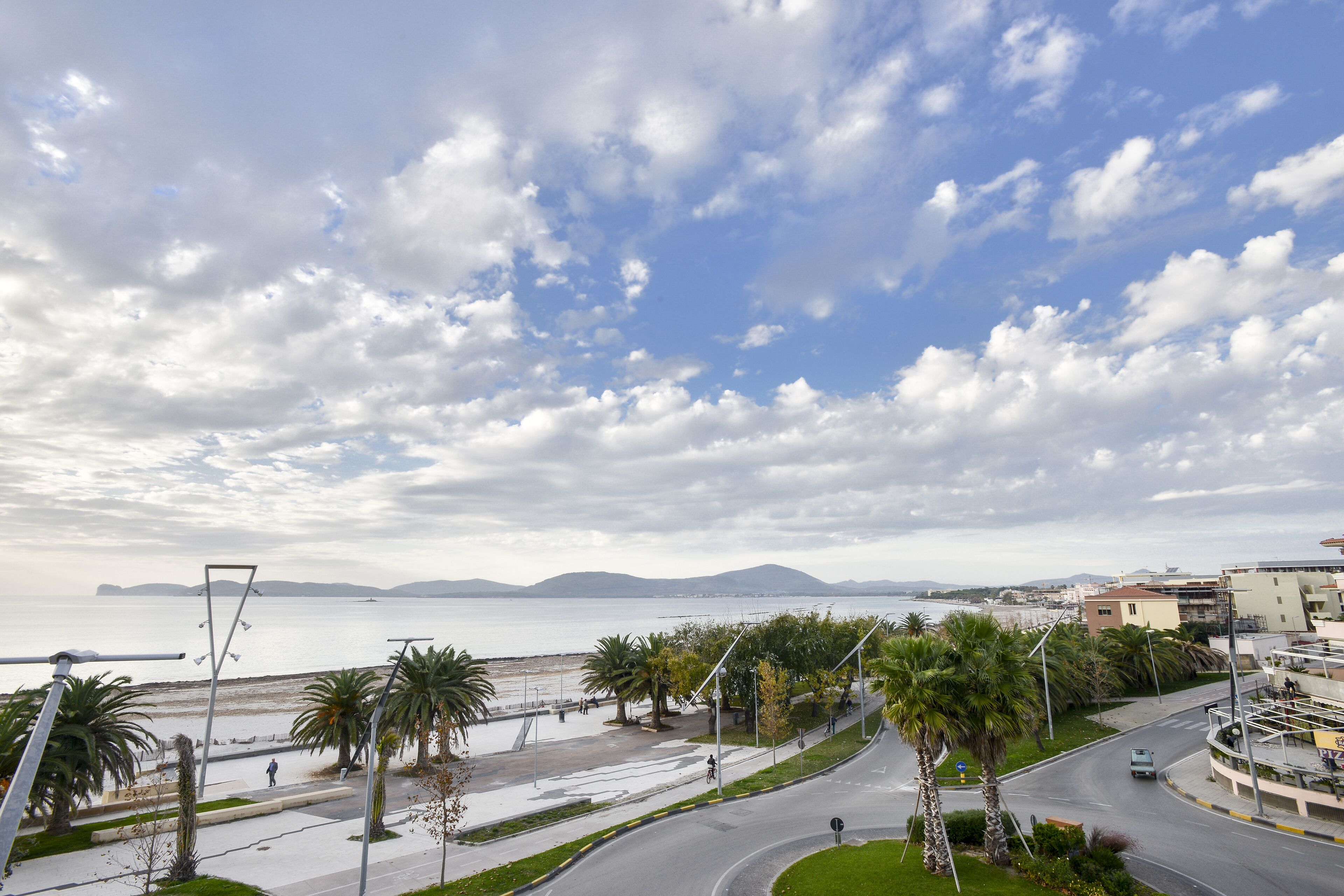 Un paisaje urbano costero con un amplio paseo marítimo, palmeras y vistas al mar y montañas distantes.
