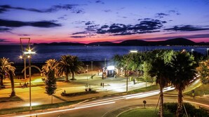 Una carretera costera con palmeras y farolas, que lleva a una zona de playa con gente y una montaña al fondo.