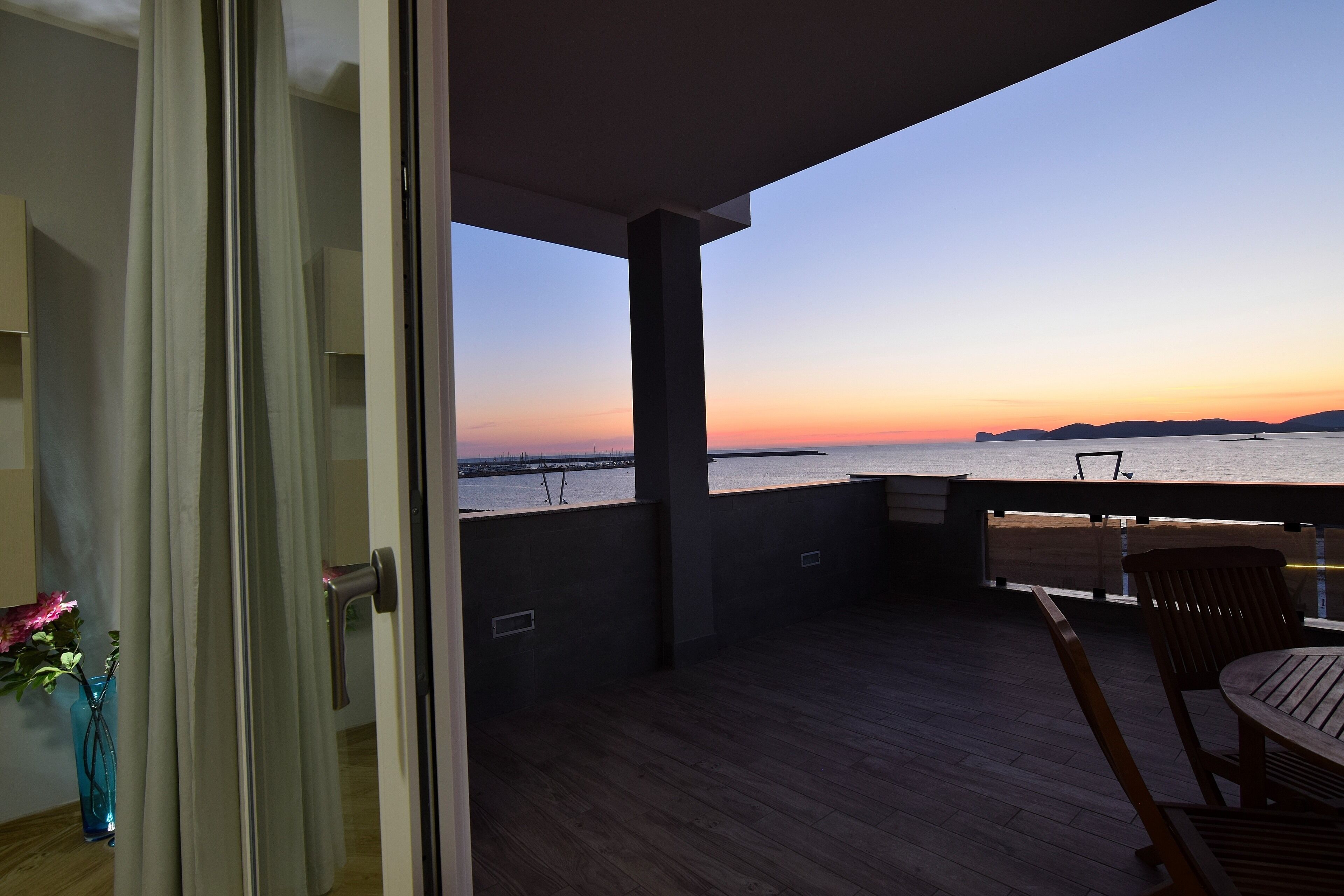 An open balcony with a view of the sunset over water, wooden flooring, and a table with chairs.