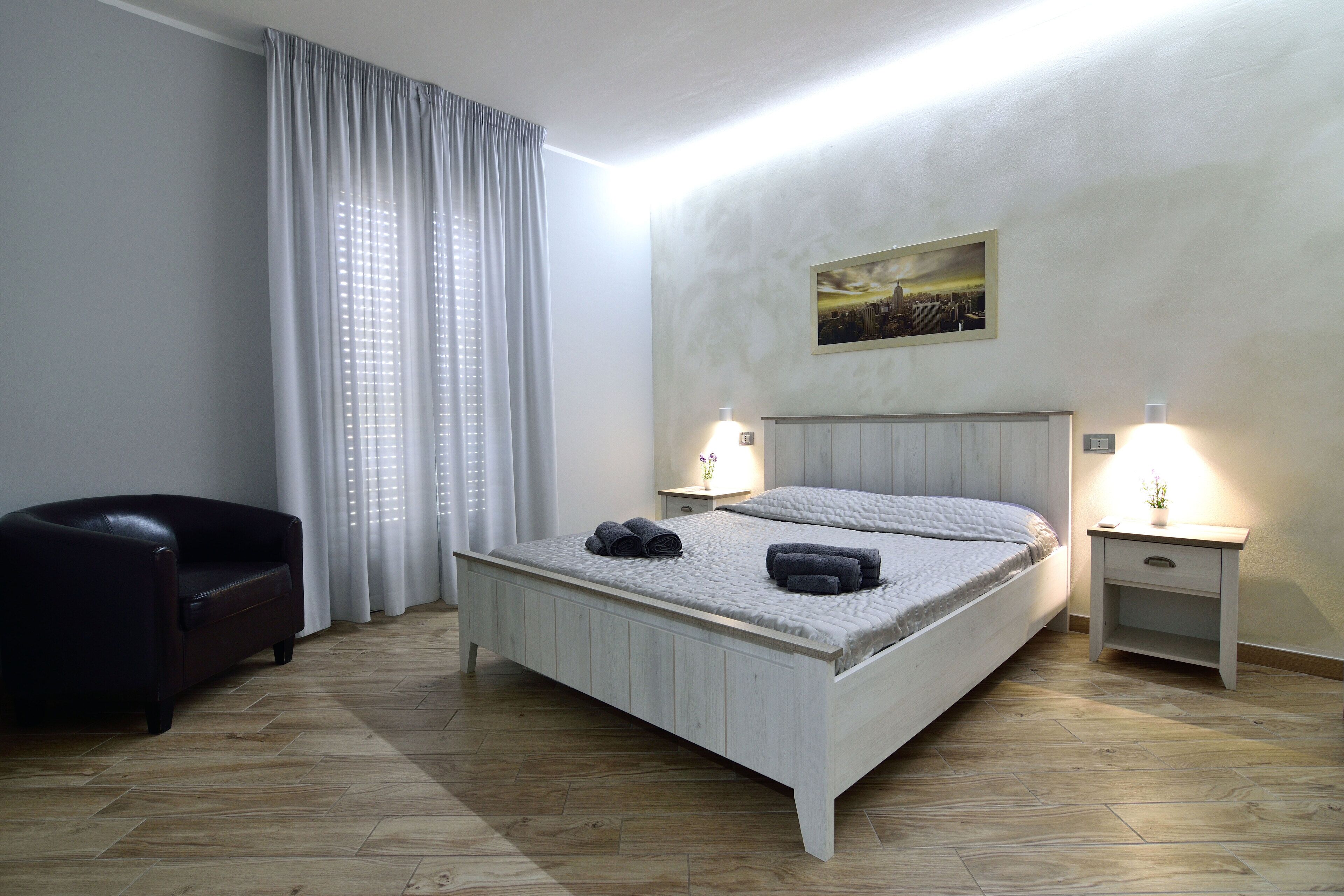 A neatly arranged bedroom with a bed, two bedside tables with lamps, a chair, and a painting on the wall.