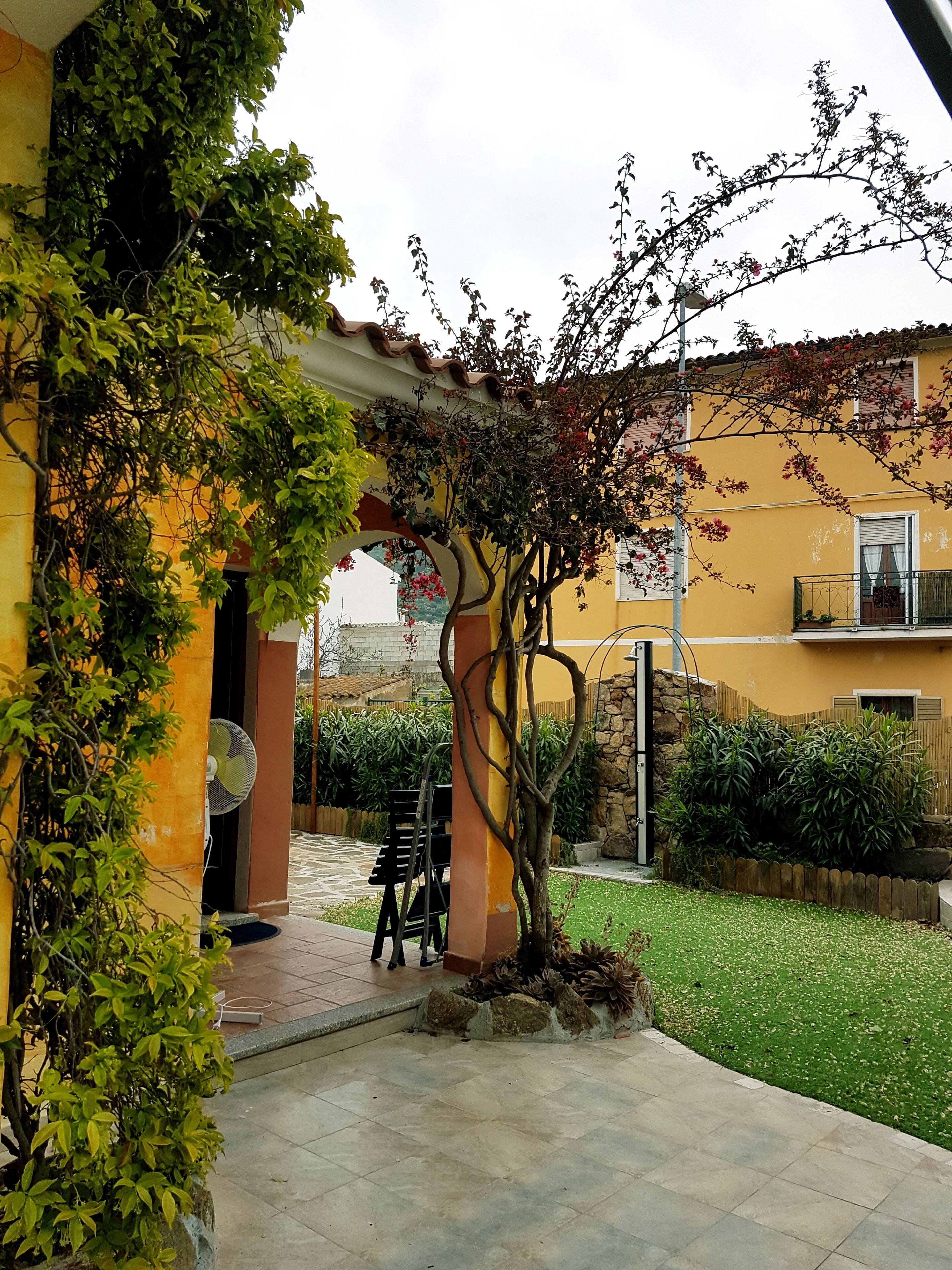 Ein Innenhof mit Fliesenbelag, einer mit Kletterpflanzen bewachsenen Pergola, einer Steinmauer und einer kleinen Sitzecke mit Stühlen.