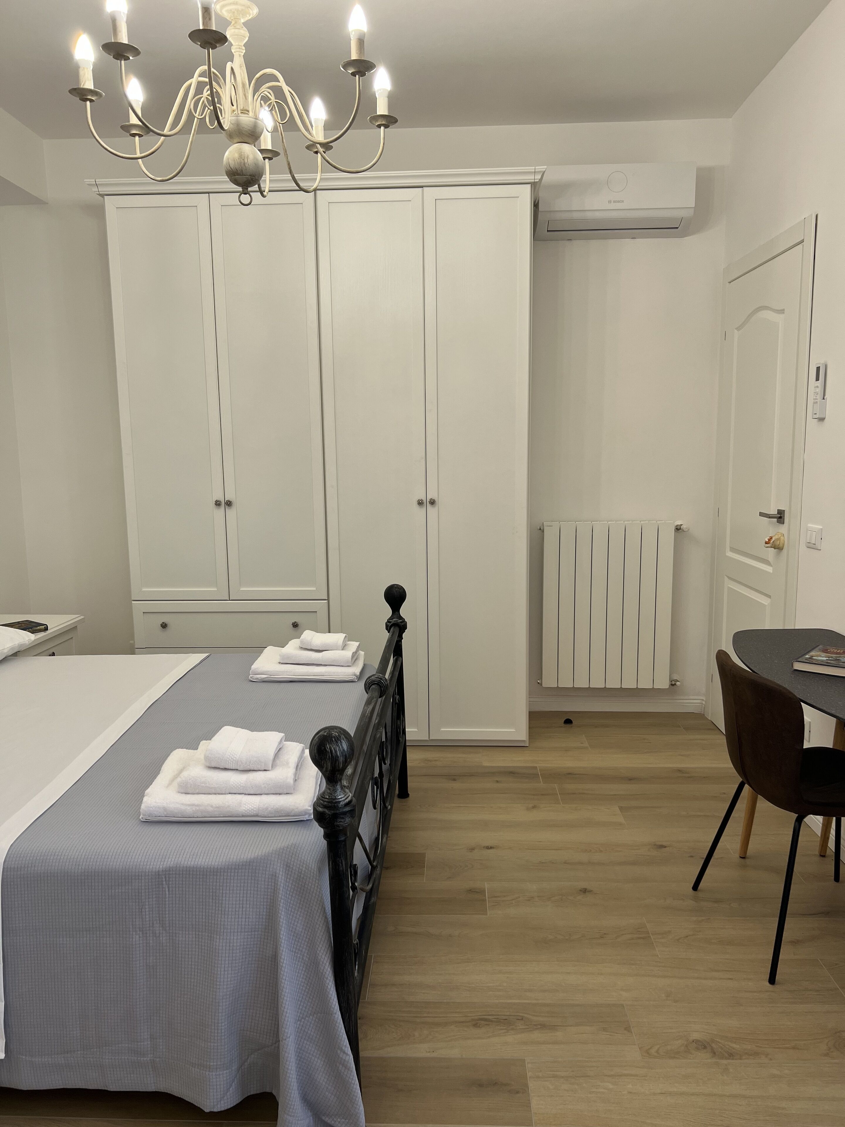 A bedroom with a bed, a chandelier, a wardrobe, a radiator, and a dining table with a chair.