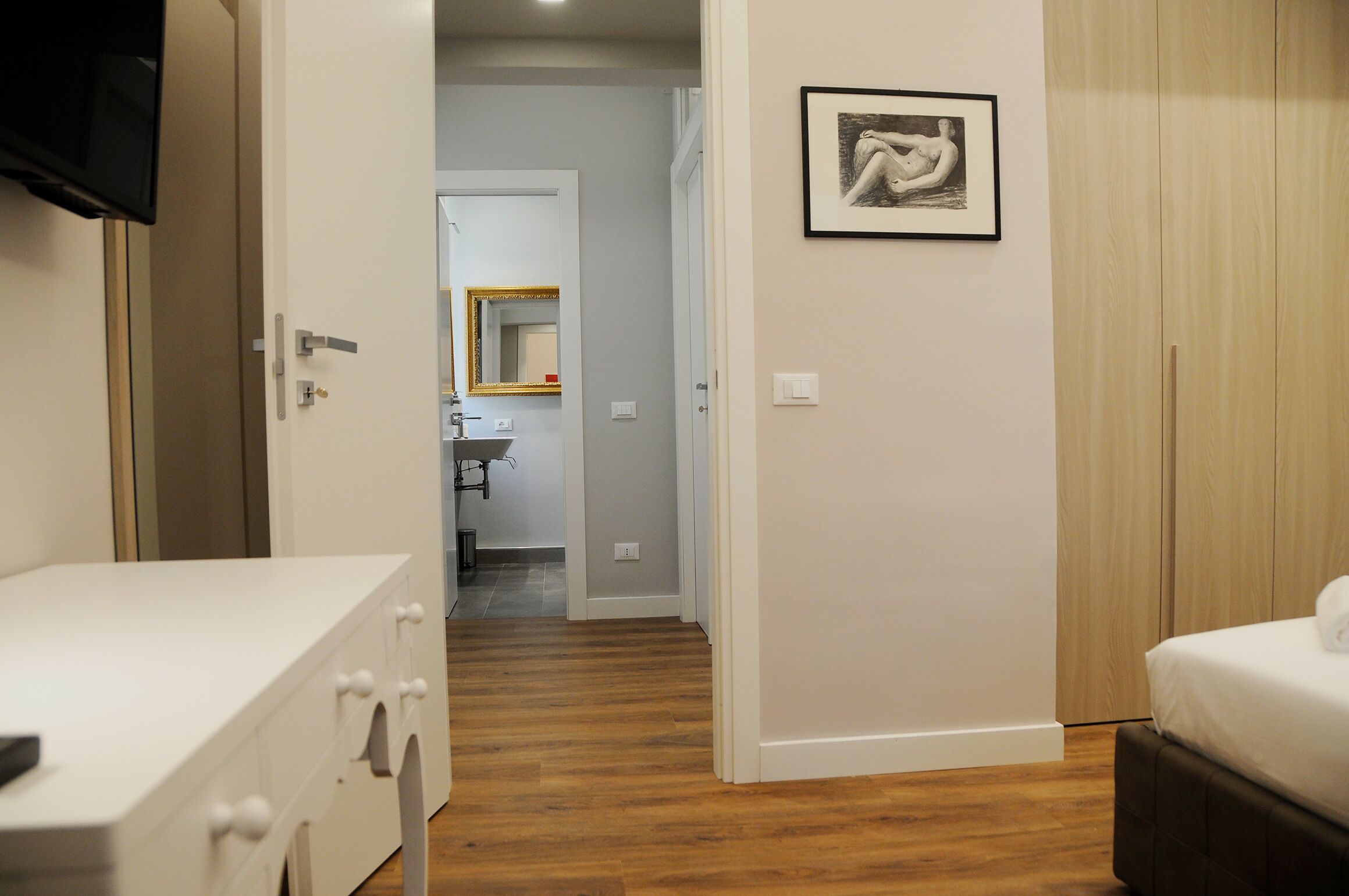 Une chambre d’hôtel avec un plancher en bois, un lit blanc, une commode blanche, un téléviseur fixé au mur et un tableau encadré accroché au mur.