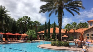 A swimming pool with lounge chairs and umbrellas, surrounded by palm trees and a building with a tiled roof.