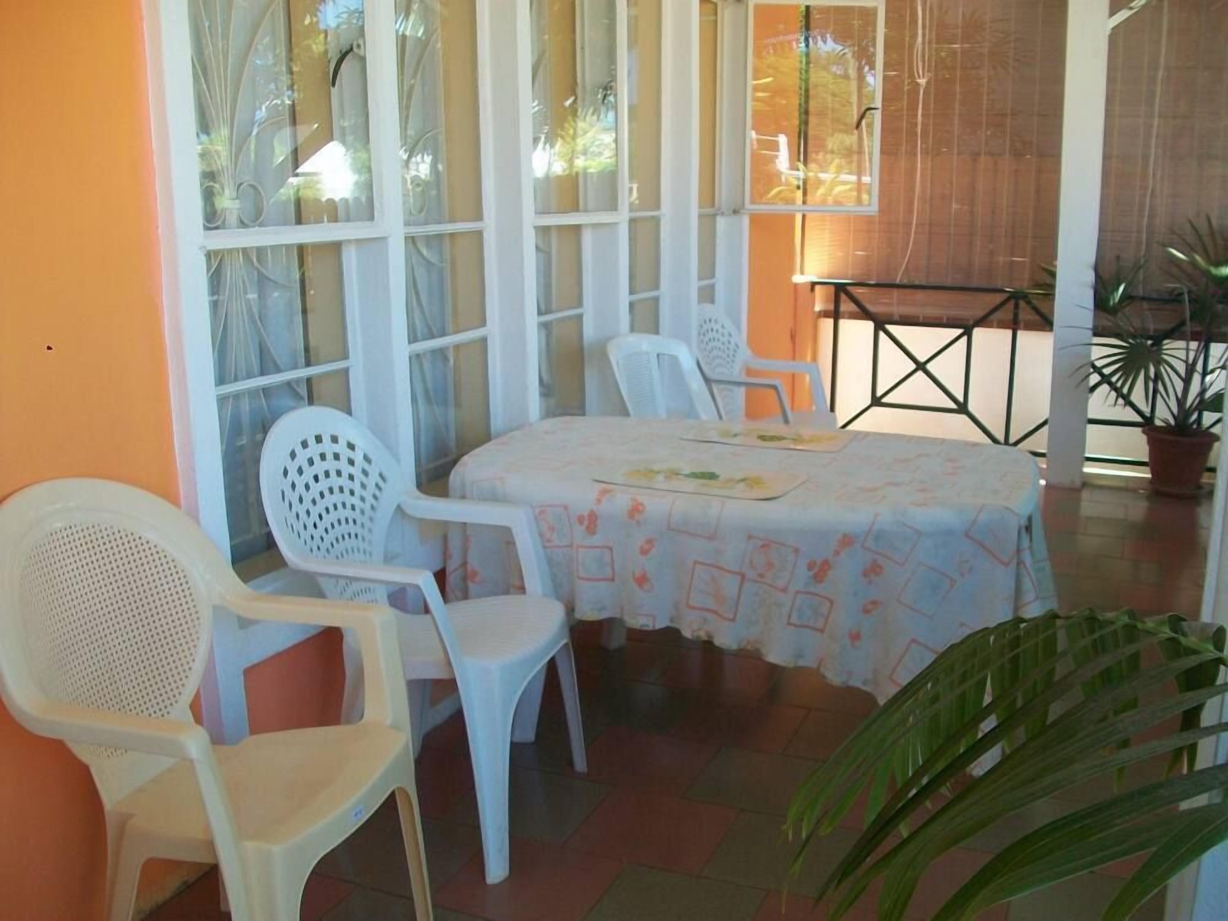 Uma área de pátio com uma mesa coberta por uma toalha de mesa florida, cercada por cadeiras de plástico branco.