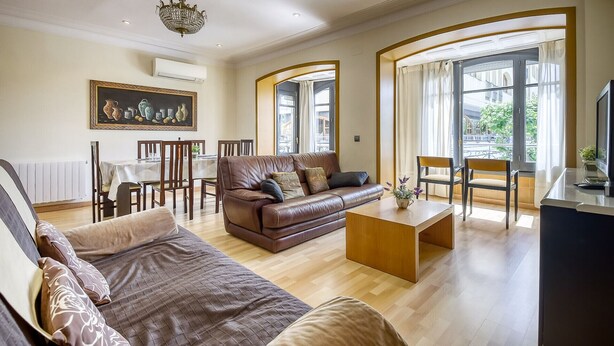 Ein geräumiges Wohnzimmer mit einer braunen Ledersofa, einem Holztisch und einem Essbereich mit Tisch und Stühlen. Ein Gemälde hängt an der Wand, und große Fenster sorgen für natürliches Licht.