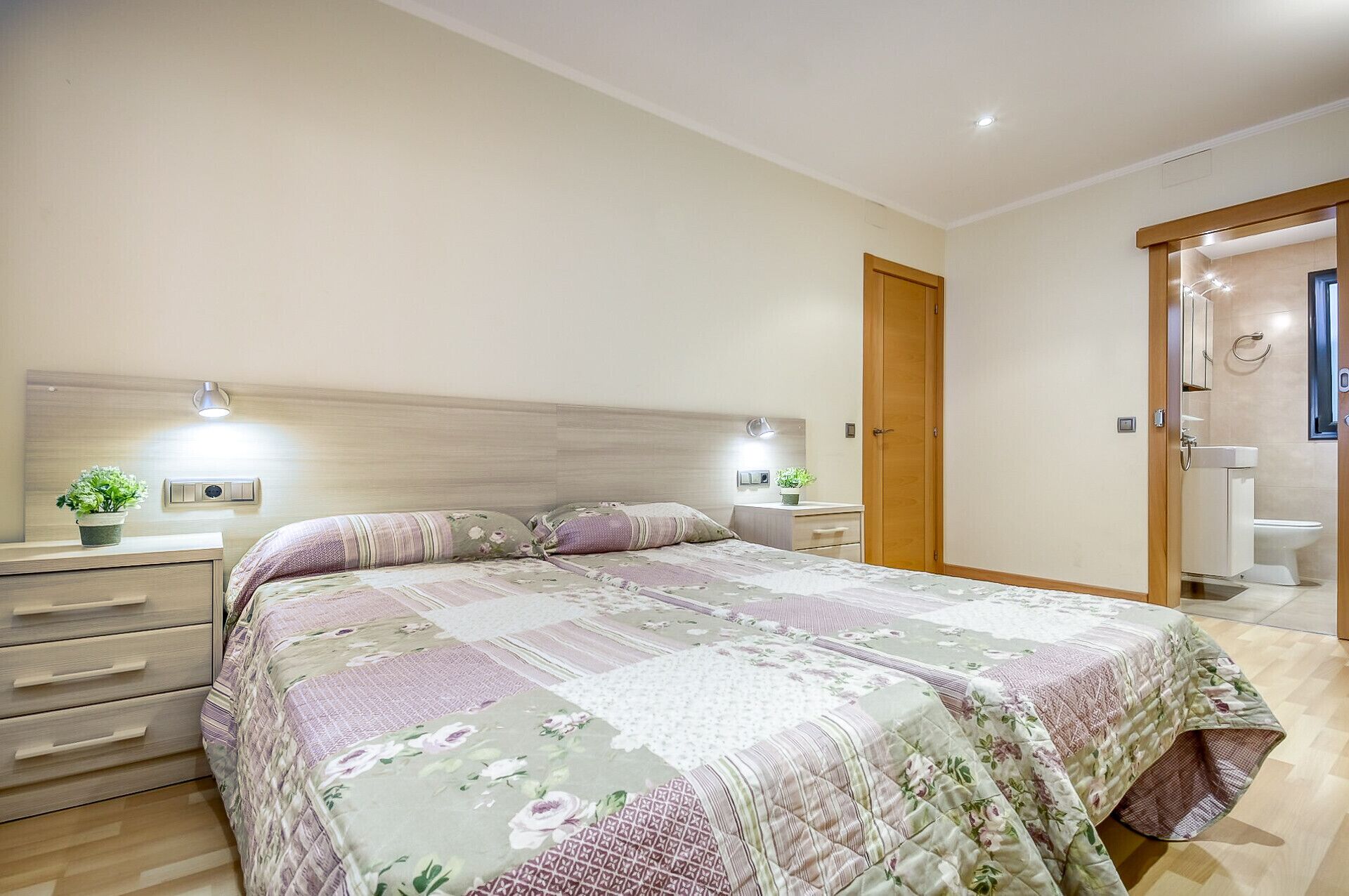 A bedroom with a bed, bedside tables, a nightstand, a wooden door, and a bathroom with a toilet and sink.