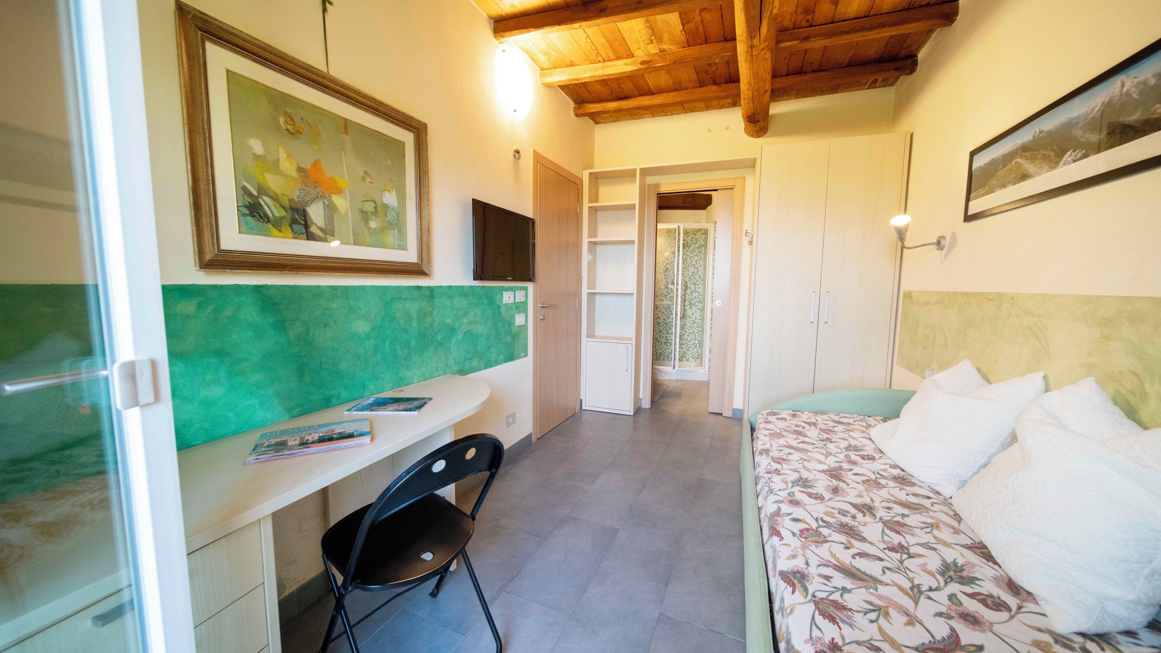 A bedroom with a bed, a desk with a chair, a painting on the wall, and a hallway leading to another room.