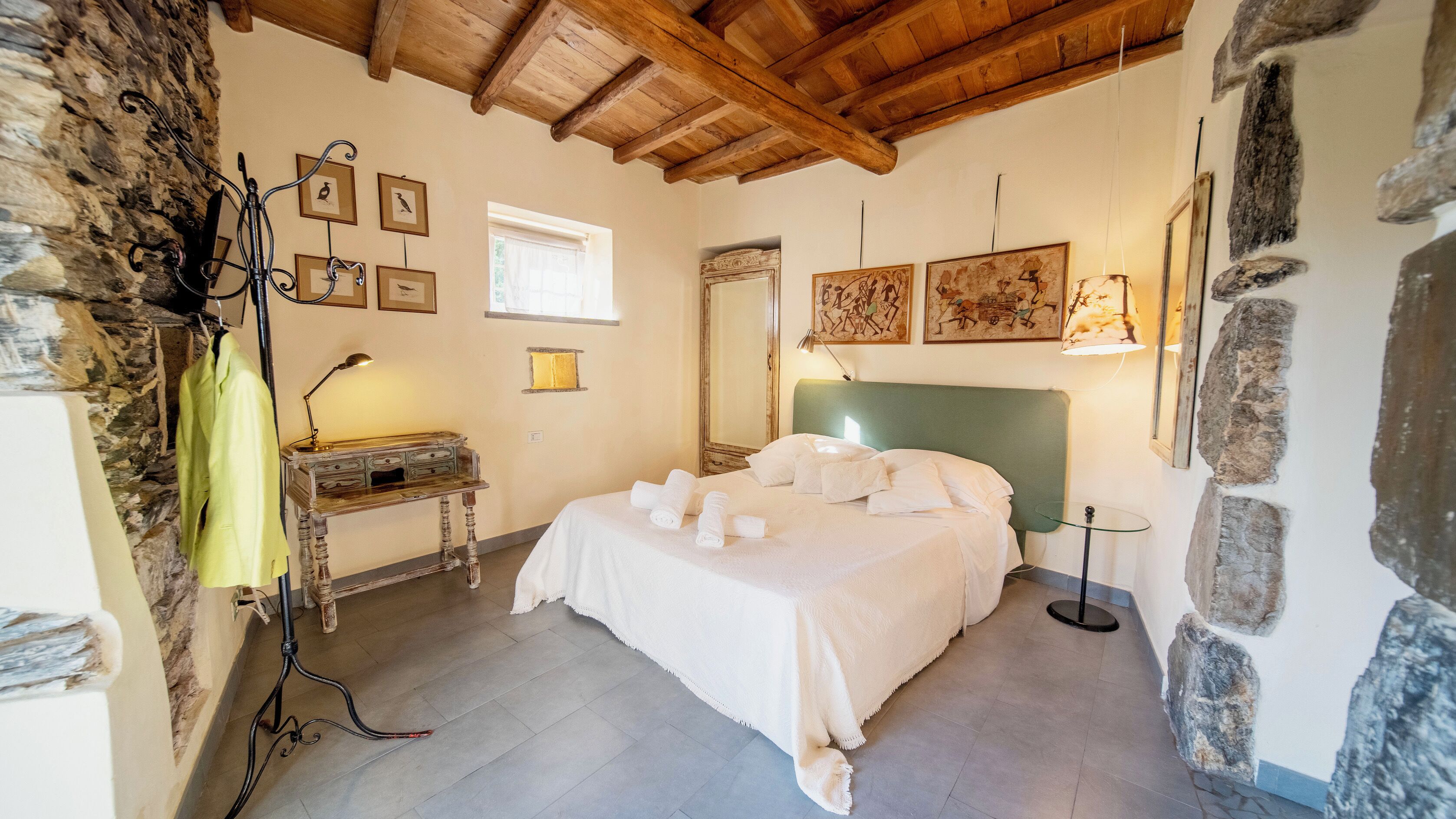 A bedroom with a stone wall, a bed with white linens, a desk with a lamp, and artwork on the walls.