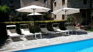 Un espace aménagé au bord de la piscine, avec des chaises longues et des parasols, une piscine en pierre et des bâtiments en pierre en arrière-plan.