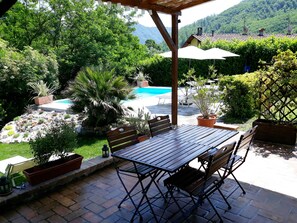 An outdoor patio with a wooden table and chairs, surrounded by potted plants and a swimming pool. - 2 Bed Modernised Barn with Pool, within easy walking distance to Town. (Bagni di Lucca)