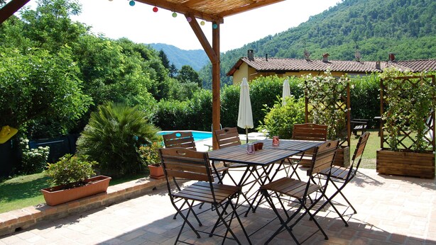 Une terrasse couverte, agrémentée d’une table et de chaises, entourée de verdure et donnant sur une piscine.