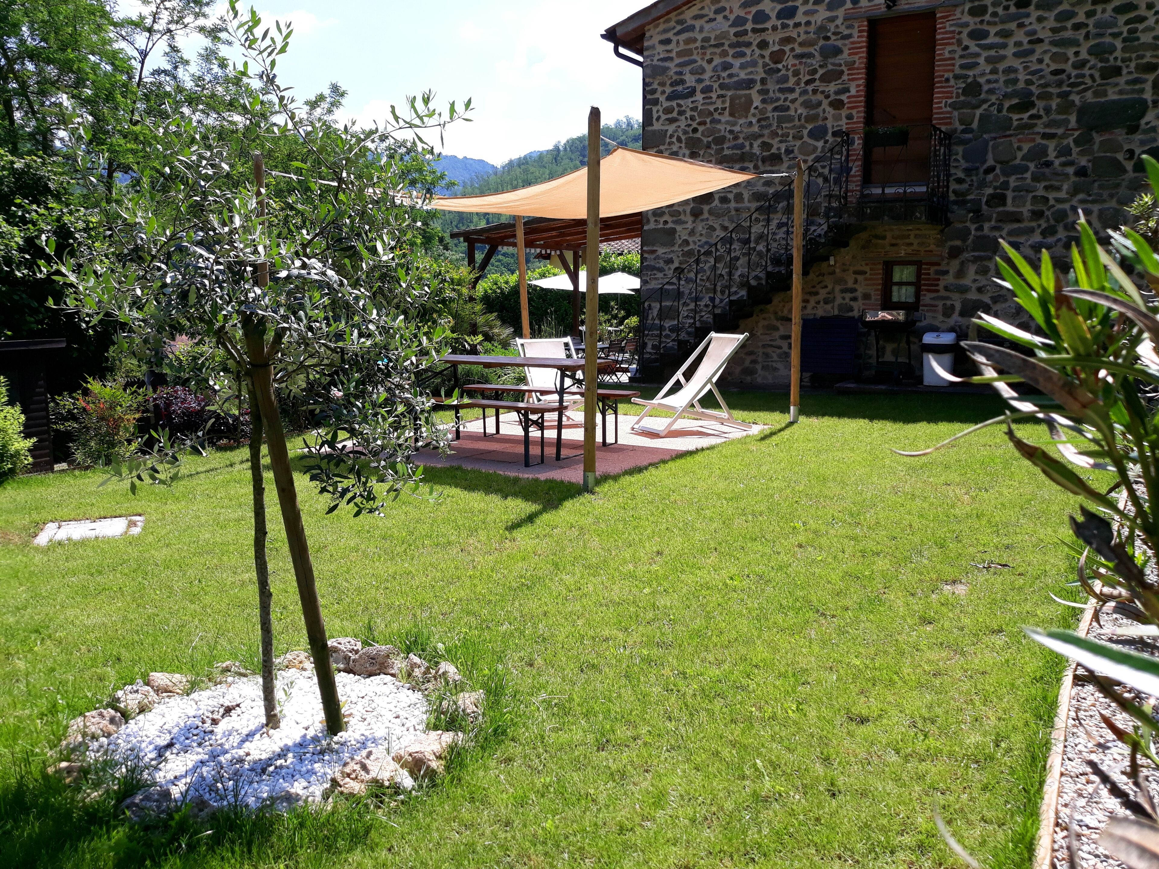 A stone building with a stone staircase, a covered patio with a table and chairs, and a grassy yard with a tree.