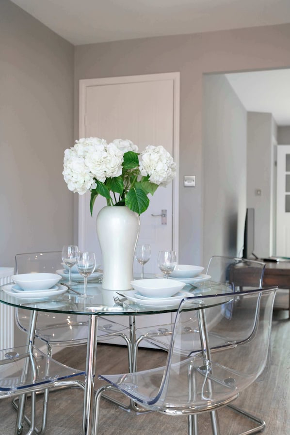 Un comedor con mesa de vidrio, sillas transparentes, platos blancos y un jarrón blanco con flores.