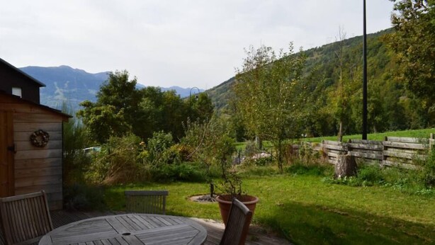 Une terrasse en bois avec une table ronde et des chaises, entourée d’un jardin luxuriant et dominant un paysage montagneux.