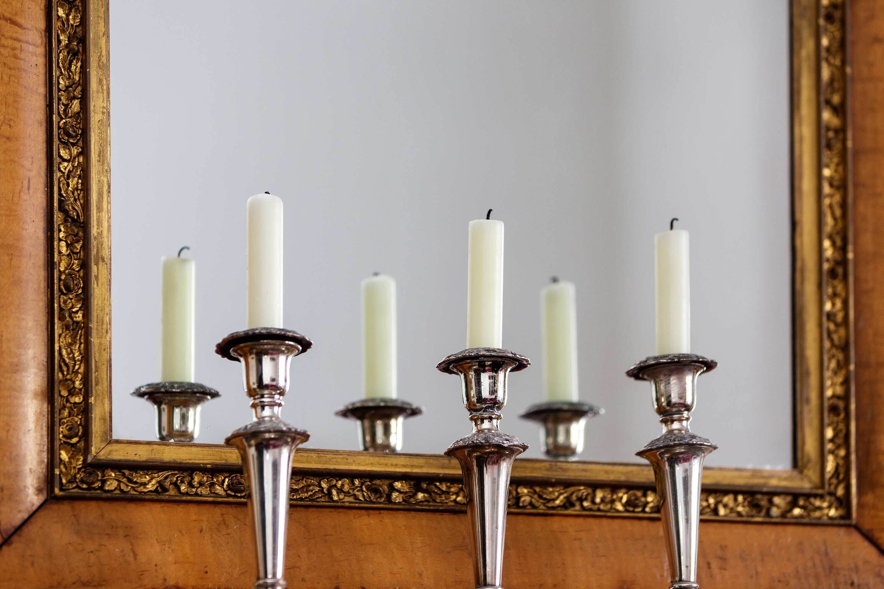 A mirror with a gold frame reflecting white candles on silver candle holders.